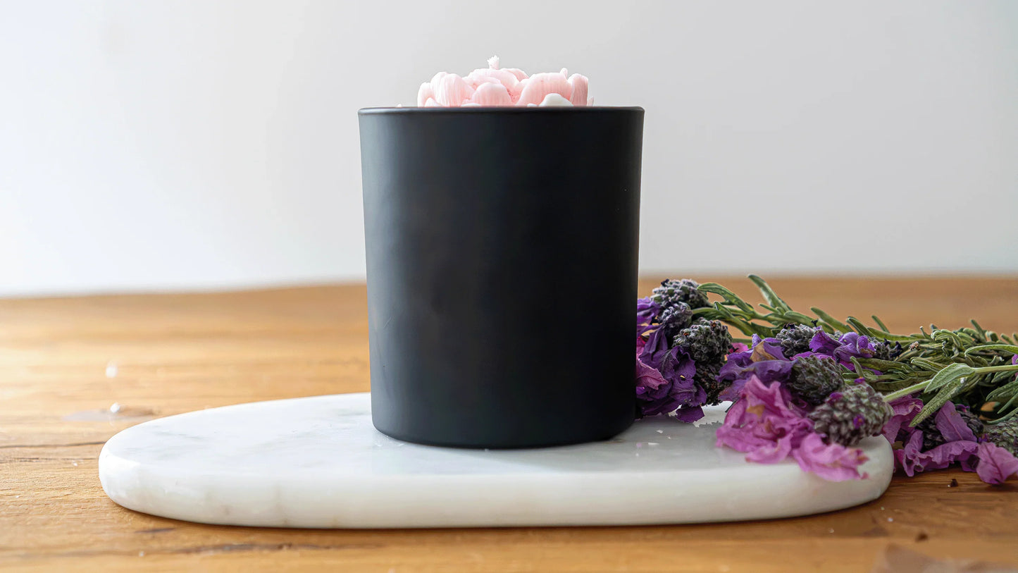 Bougie artisanale parfumée en pot noir avec motif fleur rose, posée sur plateau en marbre, fleurs lavande à côté