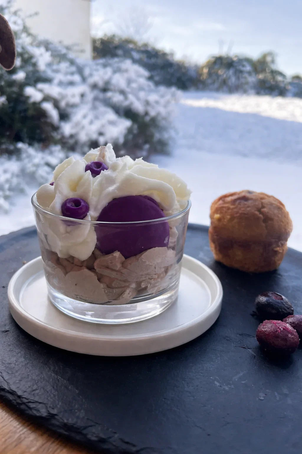 Bougie artisanale en forme de dessert gourmand avec crème et macaron violets, présentée sur ardoise, ambiance hivernale.