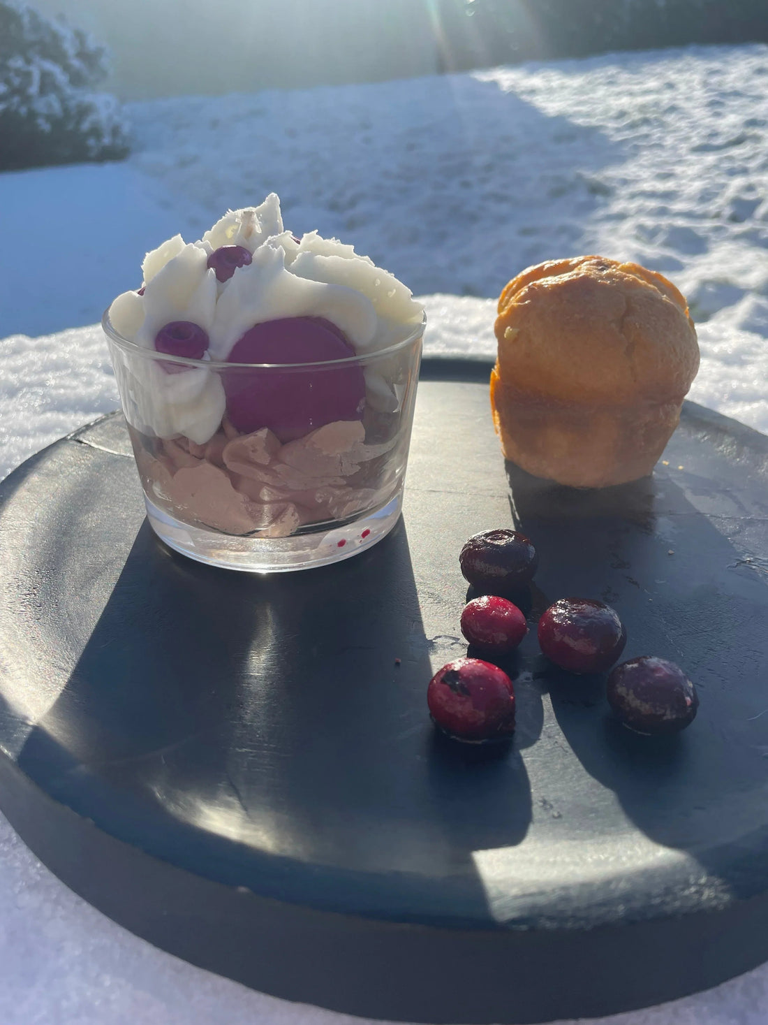 Bougie artisanale gourmande en forme de dessert avec crème et fruits rouges sur assiette noire, neige en arrière-plan.