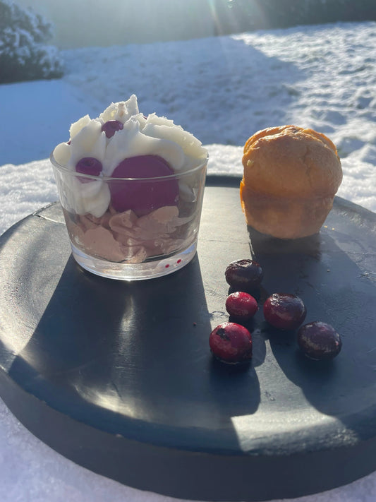 Bougie artisanale gourmande en forme de dessert avec crème et fruits rouges sur assiette noire, neige en arrière-plan.